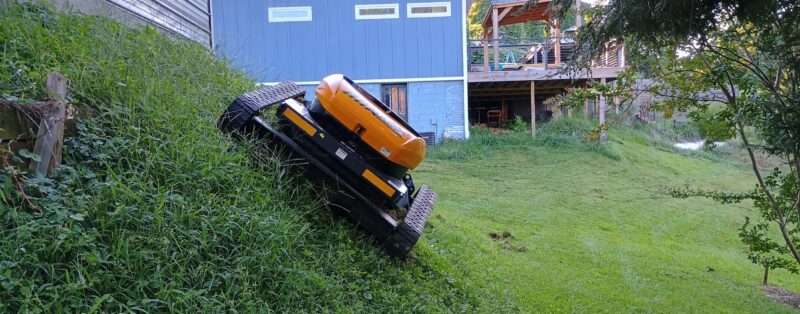 Remote-controlled slope mower cutting grass on a steep 50 degree hillside next to a residential home in Upstate South Carolina